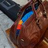 Brown leather duffel bag with clothes inside, next to a black suitcase on a concrete surface.