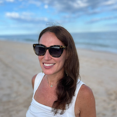 Woman wearing sunglasses and a white top on a beach with ocean and sky in the background