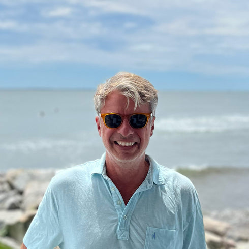 Man wearing sunglasses and a light blue shirt standing by the ocean.